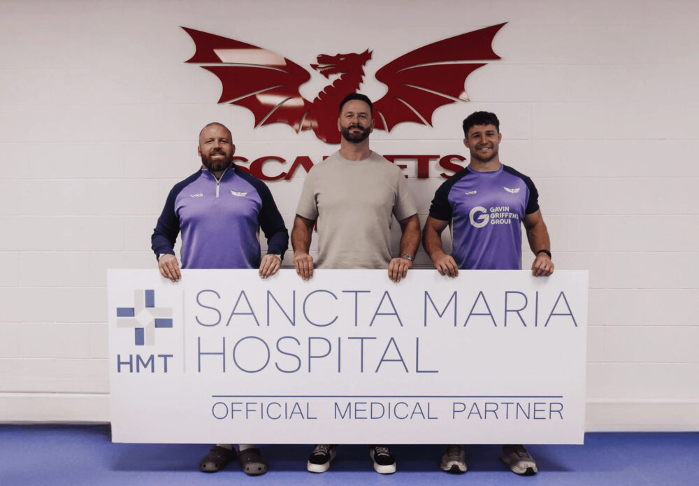 Three men hold a large HMT Sancta Maria Hospital Official Medical Partner sign beneath the red Scarlets dragon logo.