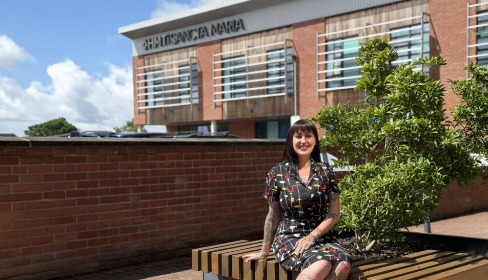 beautiful woman with tattoos wearing a rockerbilly style dress, sat on a bench outside HMT Sancta MAria hospital on a really sunny day