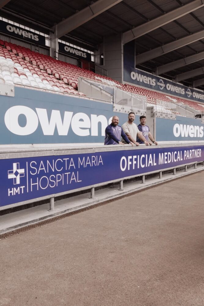 Three men lean on a pitchside barrier with an HMT Sancta Maria Hospital Official Medical Partner banner at the Scarlets stadium.